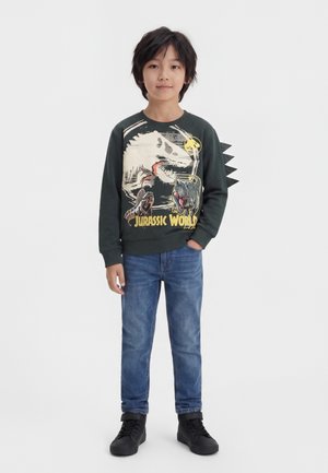 Dreng med sort hår iført mørk Jurassic World sweatshirt med dinosaurpigge på ærmerne, blå jeans og sorte sneakers, stående.
