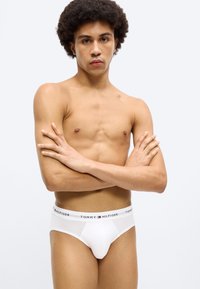 Άσπρα βαμβακερά σλιπ με μαύρη και κόκκινη ζώνη μέσης με λογότυπο. Το μοντέλο έχει ανοιχτόχρωμη επι δερμίδα και σγουρά μαλλιά, με τα χέρια σταυρωμένα στο στήθος.