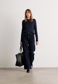 Zwarte trui en donkere jeans, gecombineerd met een zwarte tote bag. Model in elegante zwarte enkellaarsjes, staand op een lichtgekleurde ondergrond.