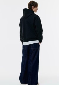 Giovane che indossa un hoodie nero, una camicia bianca e jeans larghi in denim scuro, in piedi con le mani in tasca su uno sfondo bianco semplice.