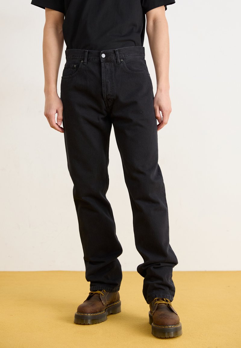 Svart denim jeans med rett passform, fem-lommers design og metallknapp lukking. Brukt sammen med brune snørestøvler på et gult gulv.
