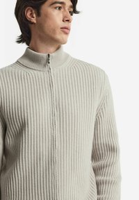 Ljusbeige tröja med ribbad textur, hög krage och hel dragkedja fram. Tyget verkar mjukt och stickat med subtila detaljer längs sömmarna.