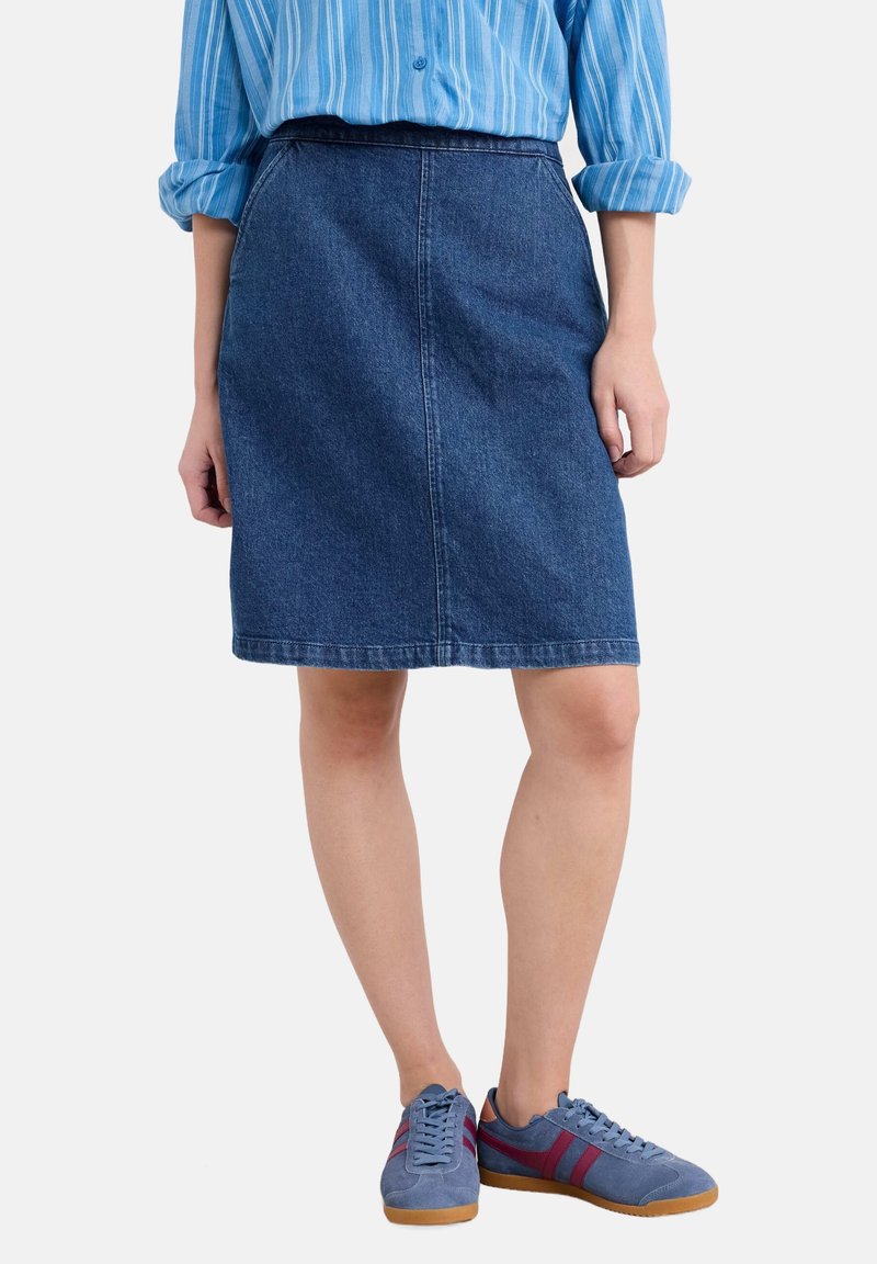 Femme portant une jupe en jean bleue, une chemise à rayures bleues boutonnée avec manches retroussées, et des baskets bleues avec des rayures rouges.