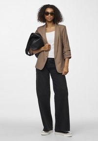 Mujer con cabello rizado que lleva un blazer marrón, top blanco, pantalones anchos negros, deportivas, gafas de sol y sostiene un bolso grande negro en el hombro.