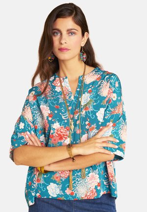 Woman with long brown hair wearing blue floral blouse, colorful earrings, gold necklaces, and bracelets, standing with arms crossed.