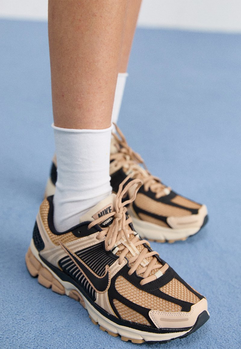 Běžecké boty Nike v béžové a černé barvě, vyrobené z síťoviny, s polstrovanou podrážkou, šněrováním v odpovídající barvě a výrazným brandingem po straně.