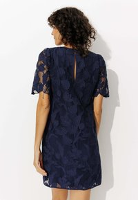 Robe en dentelle marine avec des motifs floraux, des manches courtes et un détail en trou de serrure au dos. Positionnée au-dessus du genou avec une finition texturée.
