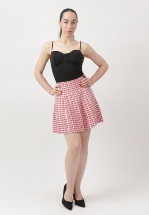 Mujer de pie con las manos en las caderas, vestida con una blusa negra sin mangas, falda a cuadros rojos y blancos, y tacones altos negros.