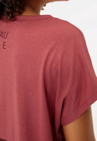 Camiseta de manga corta en color burdeos hecha de una tela suave; presenta un cuello redondeado y un texto gráfico impreso en la parte posterior.