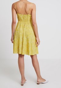 Gul blommig klänning med smockad baksida, tunna axelremmar och utsvängd kjol. Vita slip-on mules fulländar looken.