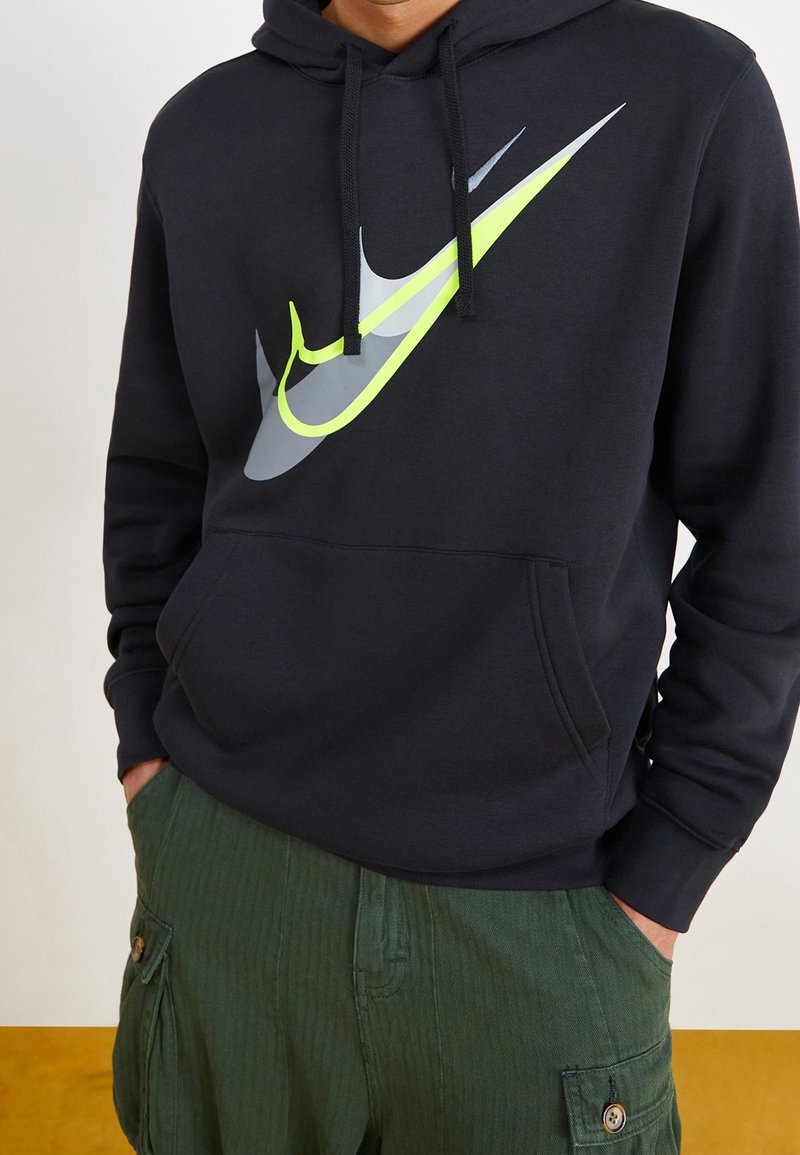 Laivastonsinisessä hupparissa suuri harmaa ja neonkeltaisen Nike-logo, jossa on etutasku ja kiristysnauhalla varustettu huppu. Yhdistetty vihreisiin cargo-housuihin.