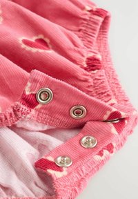 Rosa kordflord tyg med hjärtmönster. Inkluderar silverfärgade tryckknappar och draperad trim längs kanterna. Mjuk texturerad inre foder.