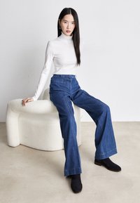 Femme portant un col roulé blanc et un jean bleu à jambes larges assise sur un banc incurvé blanc texturé sur fond uni.