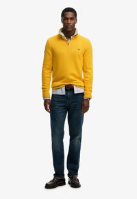 Żółty sweter dzianinowy z zamkiem na kołnierzu, noszony na niebieskiej koszuli w paski, w połączeniu z ciemnymi dżinsami i brązowymi butami, o teksturowanym wzorze.