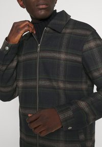 Muž v tmavě zelené a hnědé kostkované bundě s zipem přes černý pletený rolák, ruka upravující zip u límce.