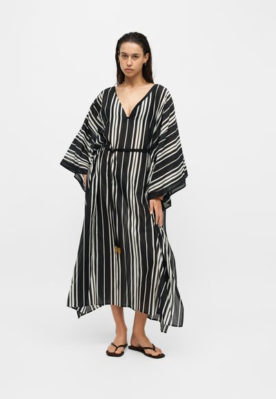 Femme portant une robe ample à rayures verticales noires et blanches, avec de larges manches et des sandales noires, debout devant un fond clair uni.