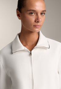 Fehér zipzáros dzseki strukturált gallérral, ezüst színű zipzárral és finom fekete díszítéssel a gallér mentén. Sima, textúrált anyag.