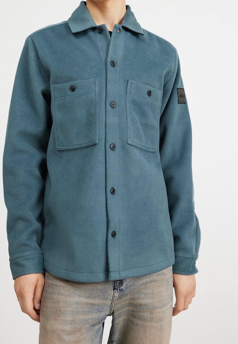 Homme portant une chemise polaire boutonnée bleu sarcelle avec deux poches poitrine et un jean bleu délavé, debout devant un fond blanc.