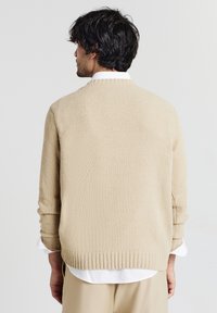 Beige gestrickter Pullover mit strukturierter Oberfläche, Rippbund und Rippärmeln sowie entspannter Passform. Darüber getragen ist ein weißes Hemd mit Kragen.