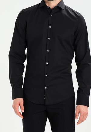 Mann trägt figurnahes schwarzes Langarm-Hemd mit Knöpfen und verbreitetem Kragen sowie schwarze Hose vor weißem Hintergrund.