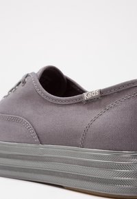 Baskets en toile grise avec semelle en caoutchouc à rayures argentées et étiquette en tissu "Keds" près du talon, vues sous un angle arrière latéral.