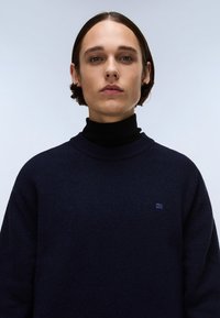 Dunkelblauer Pullover aus weichem Stoff mit hohem Kragen und gerippten Bündchen. Kleines Stoff-Logo auf der linken Brust. Glatte Textur.