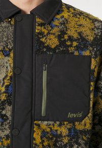 Kurtka z polaru i tkaniny z wielokolorową teksturą w żółto-niebiesko-czarne wzory, czarna kieszeń na piersi i zielony zamek błyskawiczny.