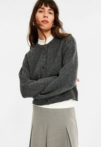 Frau mit langen braunen Haaren trägt eine dunkelgraue, geknöpfte Strickjacke über einem weißen Oberteil und einem hellgrauen, plissierten Rock vor einem weißen Hintergrund.