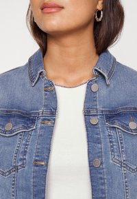 Ljusblå jeansjacka med två bröstfickor, silverfärgade knappstängningar och klassisk krage, bärs över en vit skjorta.