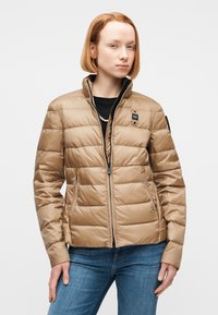 Νεαρή γυναίκα με κοντά κόκκινα μαλλιά που φοράει μια μπεζ καπιτονέ ζακέτα πάνω από μαύρο τοπ και μπλε τζιν, στέκεται μπροστά σε ουδέτερο φόντο.