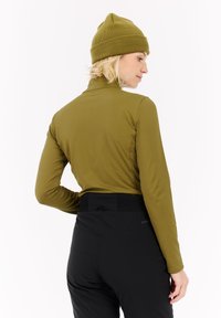 Vrouw in een olijfgroene beanie en een langetermijn top met zwarte hoge taille broek, gezien van achteren tegen een witte achtergrond.