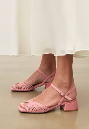 Picioare purtând sandale roz din piele întoarsă cu vârf decupat, toc gros și curele la gleznă, stând pe un podea netedă sub o rochie albă vaporoasă.