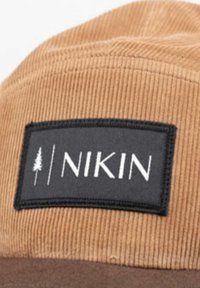 Braune Corduroy-Mütze mit einem schwarzen rechteckigen Patch, auf dem in weißer Schrift "NIKIN" und ein Baum-Logo zu sehen sind, das Materialien und Designdetails hervorhebt.