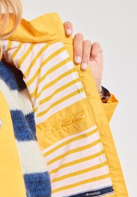 Veste de pluie jaune vif avec doublure en tissu rayé. Dispose d'une poche et d'un label "Armor Lux" au niveau de l'ourlet. Matériau imperméable.
