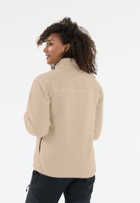 Beige fleecejacka med hög krage, texturerad yta och dragkedjefickor. Bakifrån syns ett slätt övre panel och en lös passform.