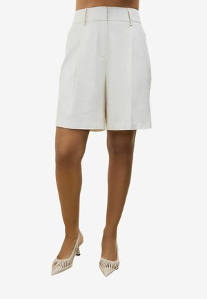 Shorts blancs sur mesure taille haute avec plis, portés par une personne à la carnation moyenne, assortis à des talons beiges à bout pointu avec détails découpés.