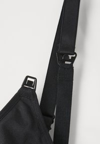 Tuvumā melna regulējama auduma siksna ar plastmasas sprādzi un āķi uz gaiša fona.