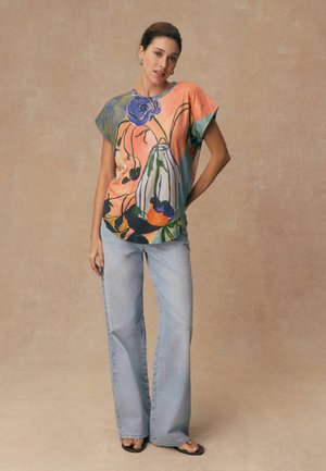 Kvinna som står med ena handen på höften, klädd i en färgglad topp med blommönster, ljusblå vida jeans och svarta sandaler mot en beige bakgrund.