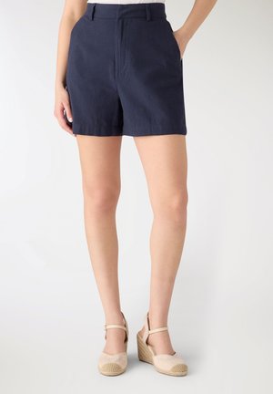 Donna che indossa pantaloncini blu navy a vita alta e espadrillas beige con zeppa, in piedi con una mano in tasca davanti a uno sfondo semplice.