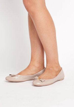 Ben klædt i blanke nude balletflade sko med små dekorerede sløjfer og guld firkantede detaljer på en hvid baggrund.