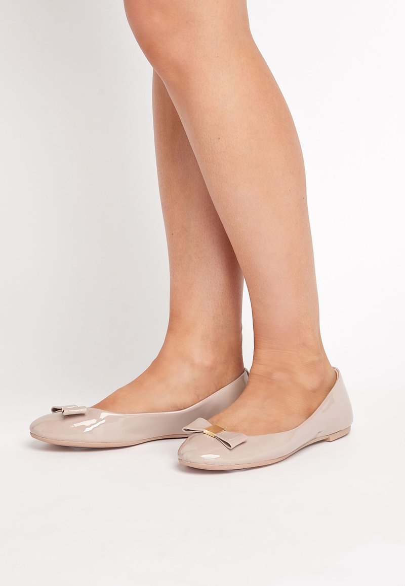 Jambes portant des ballerines nues brillantes avec de petits nœuds décoratifs et des accents carrés dorés sur un fond blanc.
