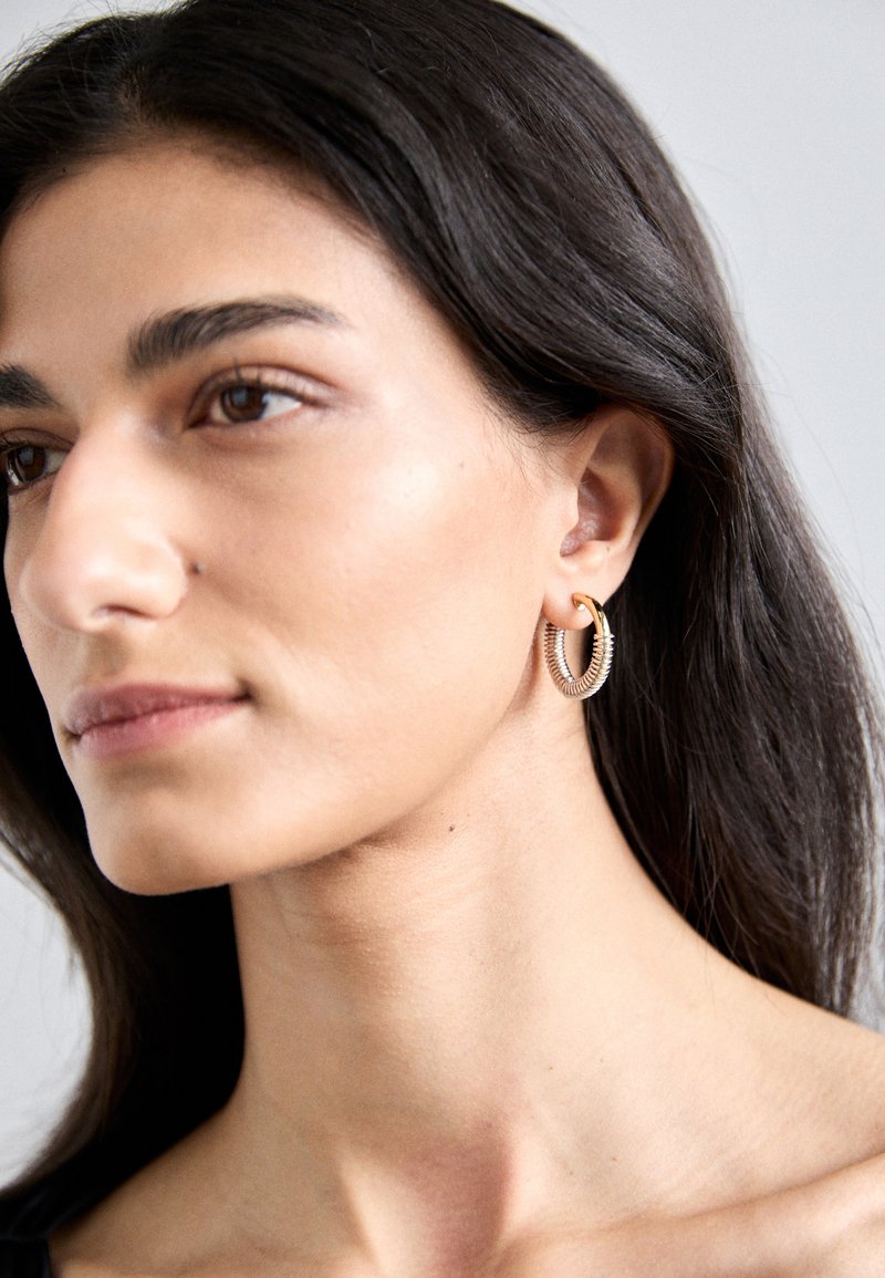 Χρυσά και ασημένια σκουλαρίκια κρίκοι με σπειροειδή σχέδιο και ραβδωτή υφή, κρεμαστά από μια μικρή χρυσή λεπτομέρεια. Ο τόνος του δέρματος και το χρώμα των μαλλιών είναι ουδέτερα.