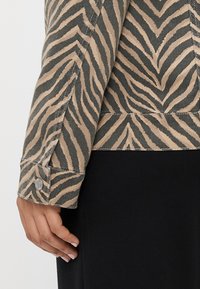 Zebraprint denimjakke med beige og mørkegrønne striber, der har en knaplukning på ærmet og tekstureret stof. Nærbillede af ærmet og hånden.