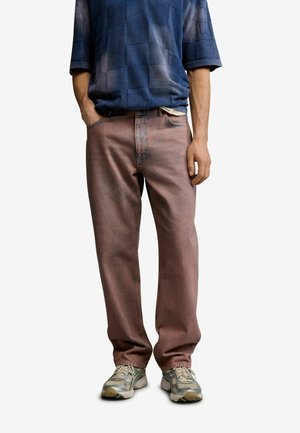 Voksen iført falmede lyserøde jeans med lige ben, blå ternet kortærmet skjorte og beige atletiksneakers, stående med hænderne afslappet.