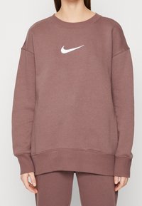 Sudadera de gran tamaño en color malva, hecha de tela suave, con cuello redondo, puños y dobladillo acanalados, y un logotipo de Nike en blanco en el pecho.