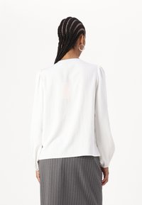 Blusa bianca con maniche lunghe a sbuffo, vestibilità ampia e collo rotondo, abbinata a una gonna grigia a righe sottili. Texture liscia, design minimalista.