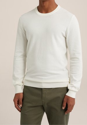 Man draagt een aansluitende, lange mouwen, witte trui met ronde hals en olijfgroene geruite broek, staand tegen een beige achtergrond.