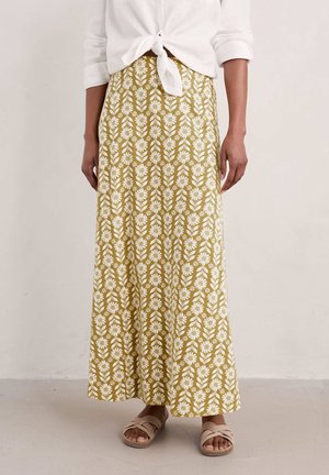 Kvinde iført en gul maxinederdel med hvidt blomsterprint, en hvid bundet bluse og beige flettede sandaler, stående på et lyst gulv.