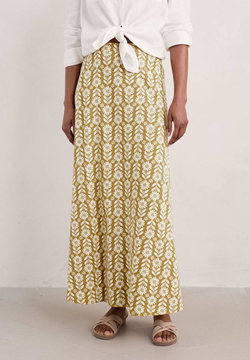 Femme portant une jupe longue jaune à imprimé floral blanc, une blouse blanche nouée et des sandales tressées beige, debout sur un sol clair.