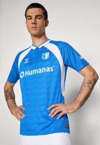 Man in een blauw-wit 1. FC Magdeburg voetbalshirt met het sponsorlogo "Humanas", staand met handen op de heupen tegen een effen achtergrond.